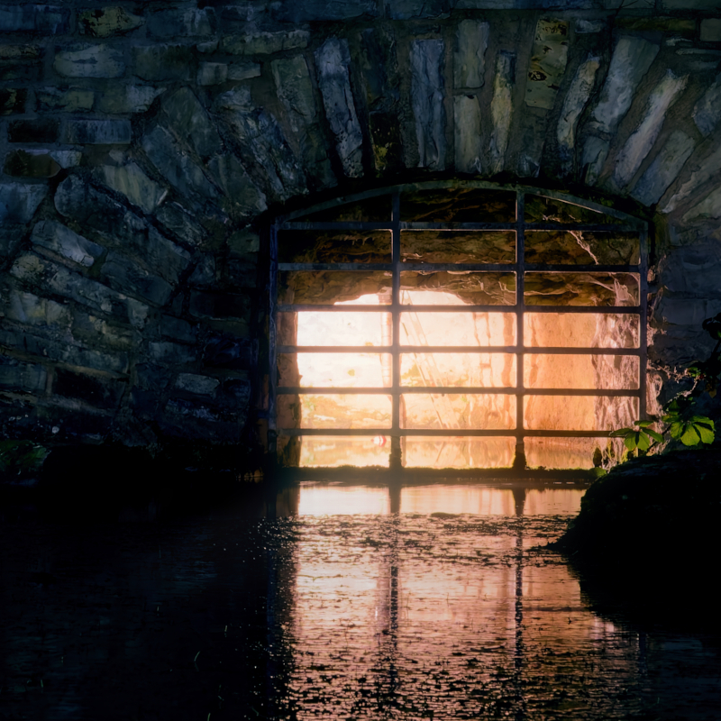 Dunkler Wasserdurchlass aus Stein mit warmem Licht hinter einem Gitter und Spiegelung im Wasser.